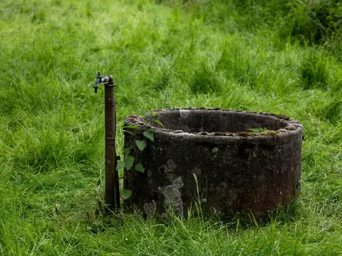 An old stone well with a metal tap in long green grass