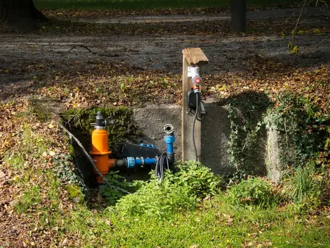 A well pipe, filter and pump hookup set in a stone-lined pit at the edge of a sunny lawn