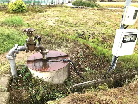 A covered wellhead with its pipe and valve and a pump control box in a grassy yard